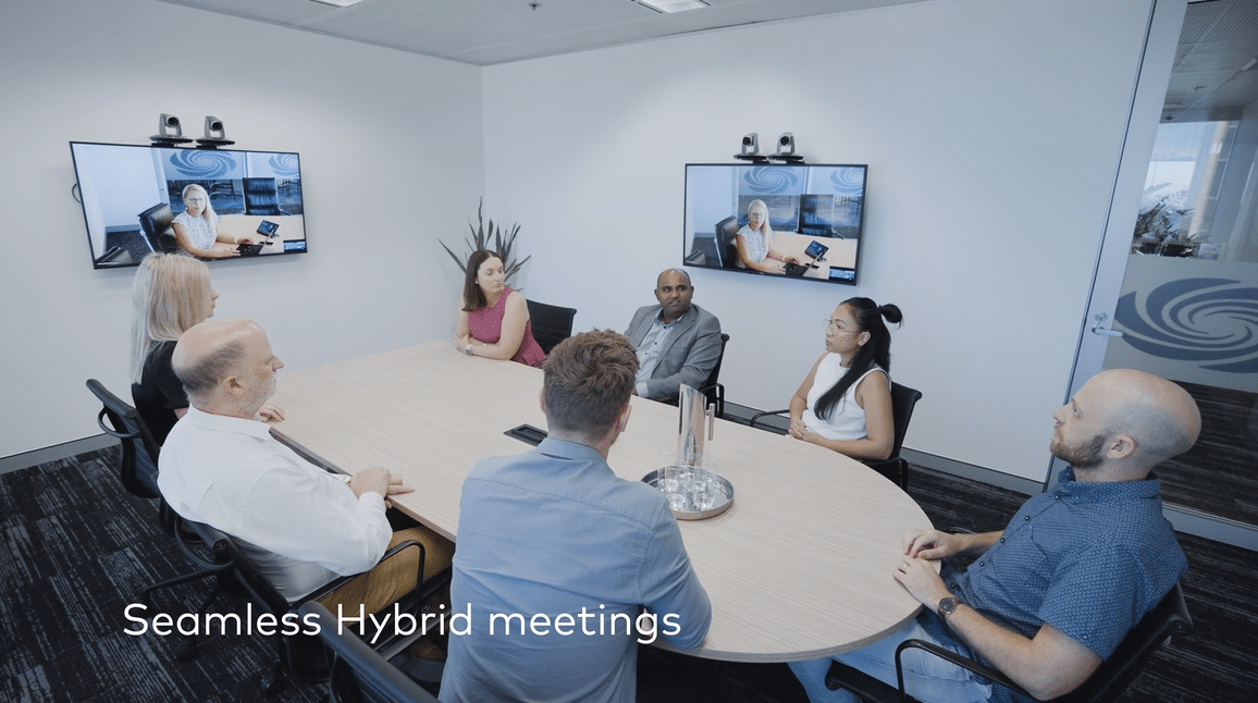 Seamless Hybrid Meetings: 7 people, in-person & virtual, gathered around a table with a plant backdrop. Less than 125 characters.