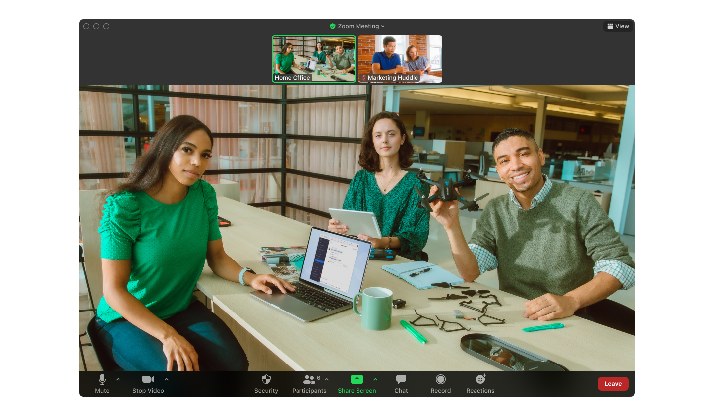 Group video call: diverse trio smiles at camera; laptop, phone, coffee, and office supplies scattered on the table, call icons visible.