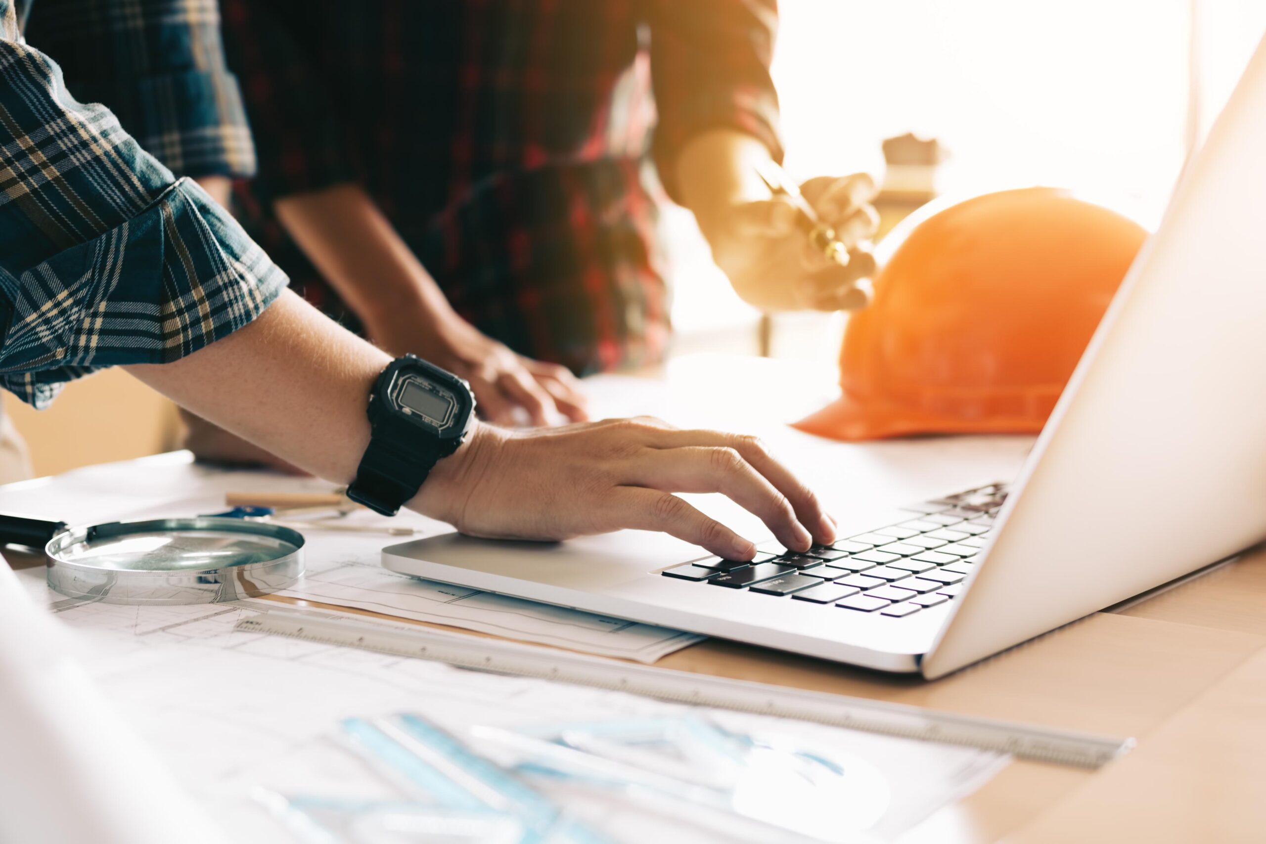 Two people collaborate at a table with a laptop, designering tools, and an orange hard hat; one types while the other points.