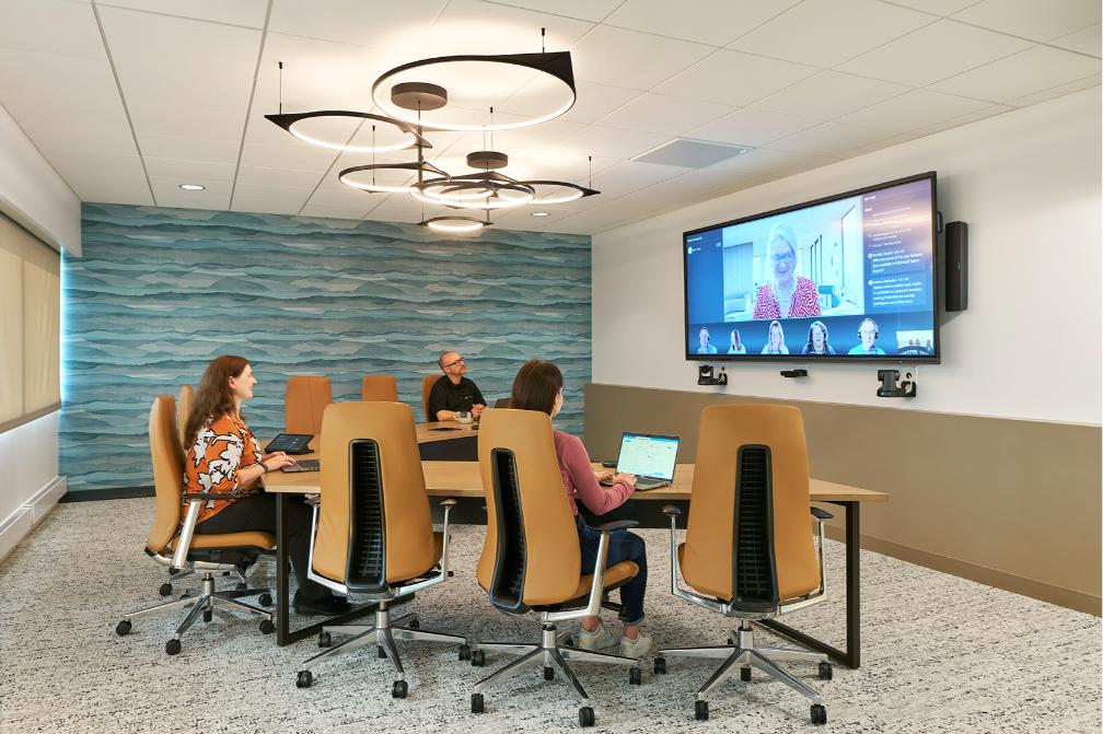 A modern conference room with three people on a video call, blue wall, brown chairs, circular lights, each with a laptop.